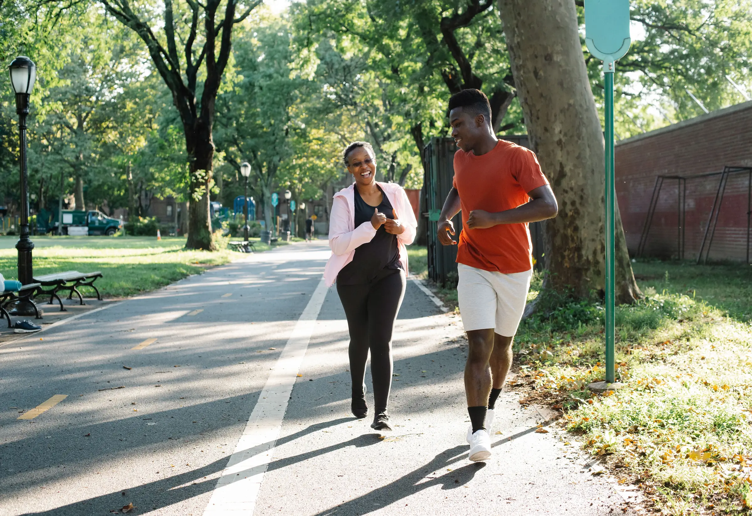 A trainer and client laughing as they jog together along a tree-lined park path