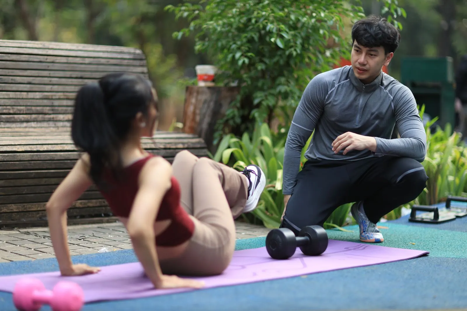A trainer kneels beside a client to guide her through a floor exercise in a park