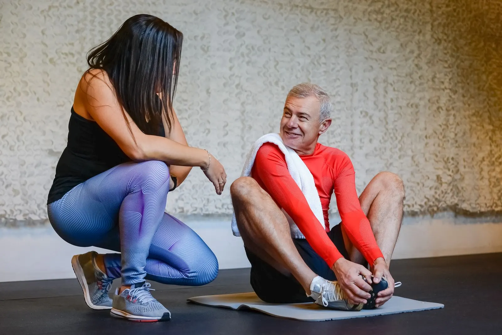 A trainer crouches to chat with a smiling client as he stretches on a mat after a workout