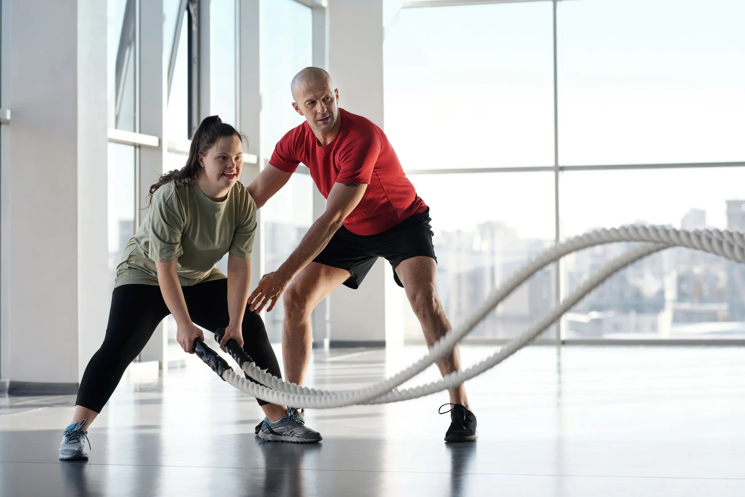 A trainer guides a smiling client through a battle rope exercise in a bright studio with city views