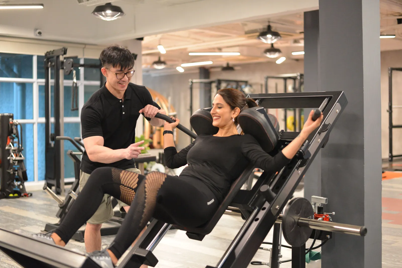 A smiling trainer guides a smiling client on a leg press machine