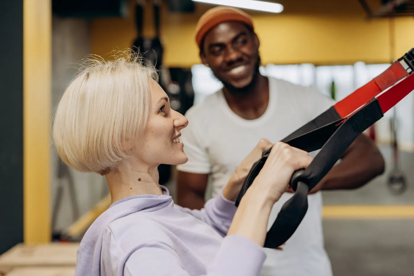 A trainer guiding a smiling woman through a suspension-strap exercise in a gym