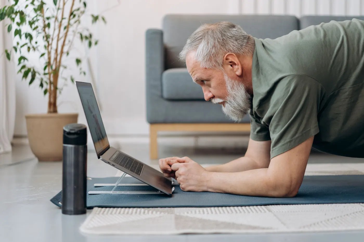 An older man holding a plank on a mat at home while following a session on his laptop