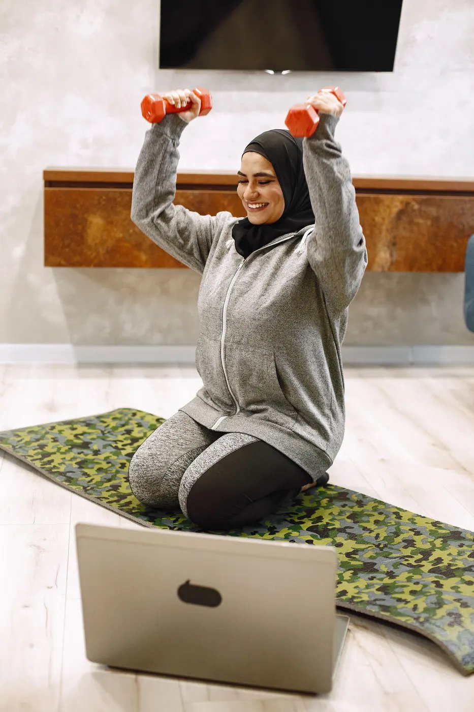 A woman in a headscarf lifting light dumbbells on a mat at home, facing her laptop