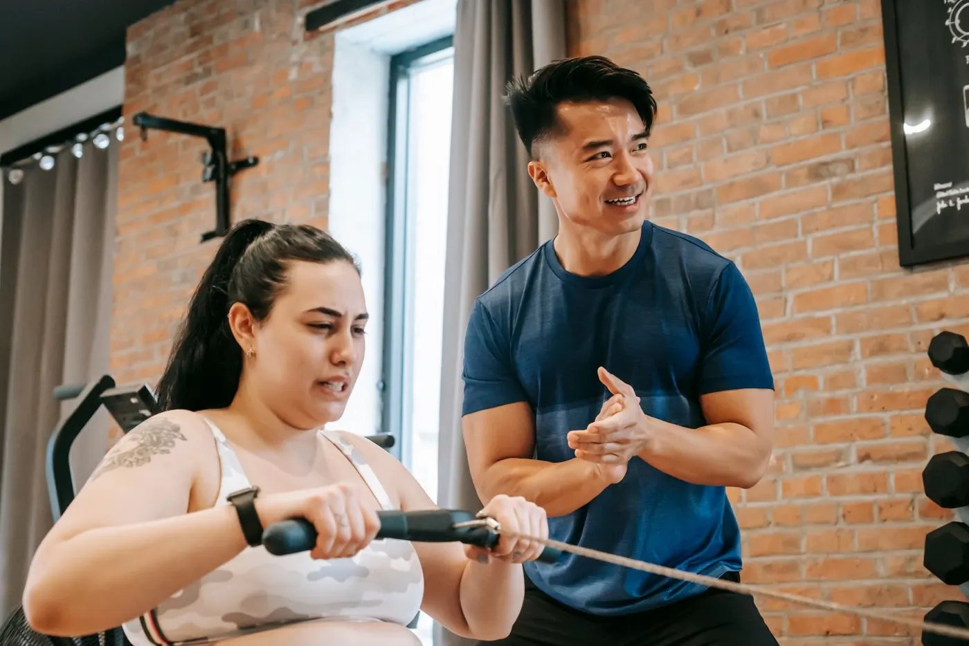 A trainer cheering on a woman as she does a cable row in a brick-walled gym