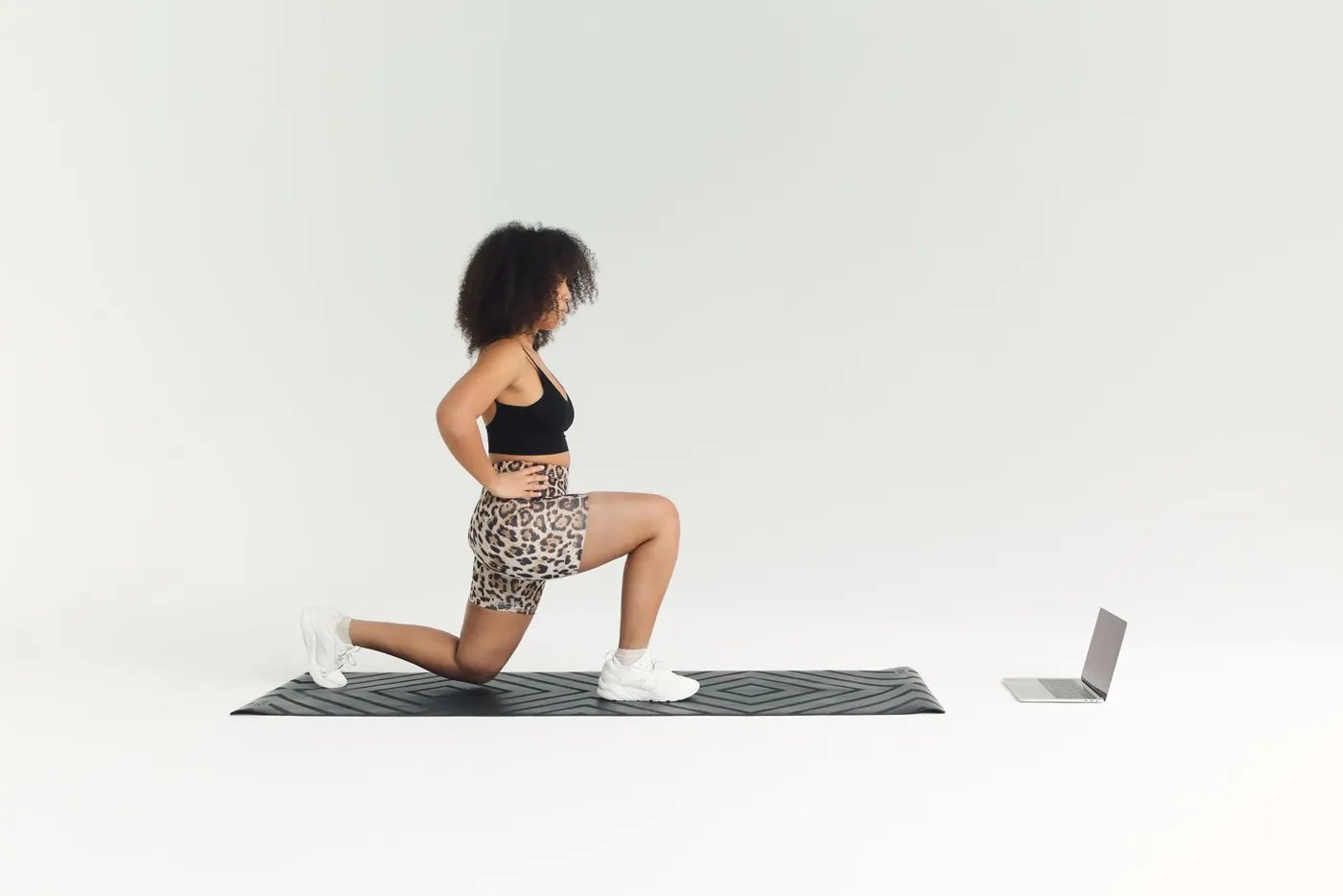 A woman doing a lunge on a mat facing an open laptop