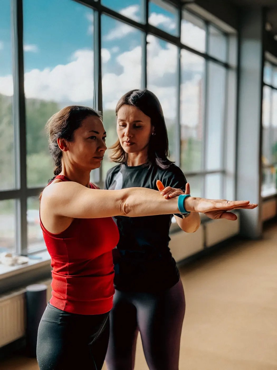 A trainer guiding a woman's outstretched arm in a bright gym with tall windows