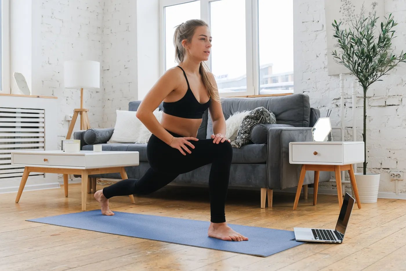 A woman in workout clothes doing a lunge on a mat in her living room, facing a laptop on the floor
