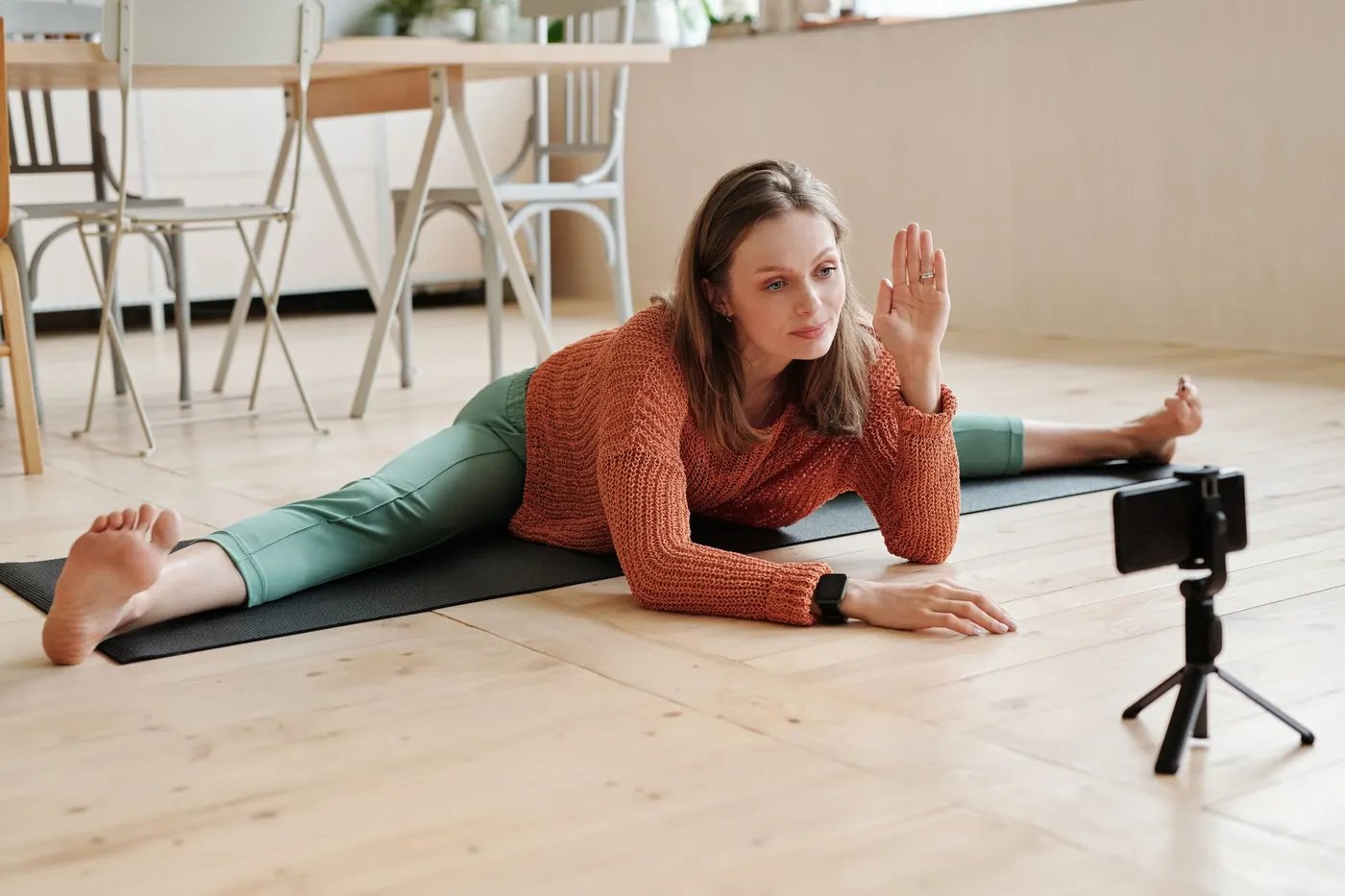 A woman stretching on a mat at home and waving at her phone on a tripod