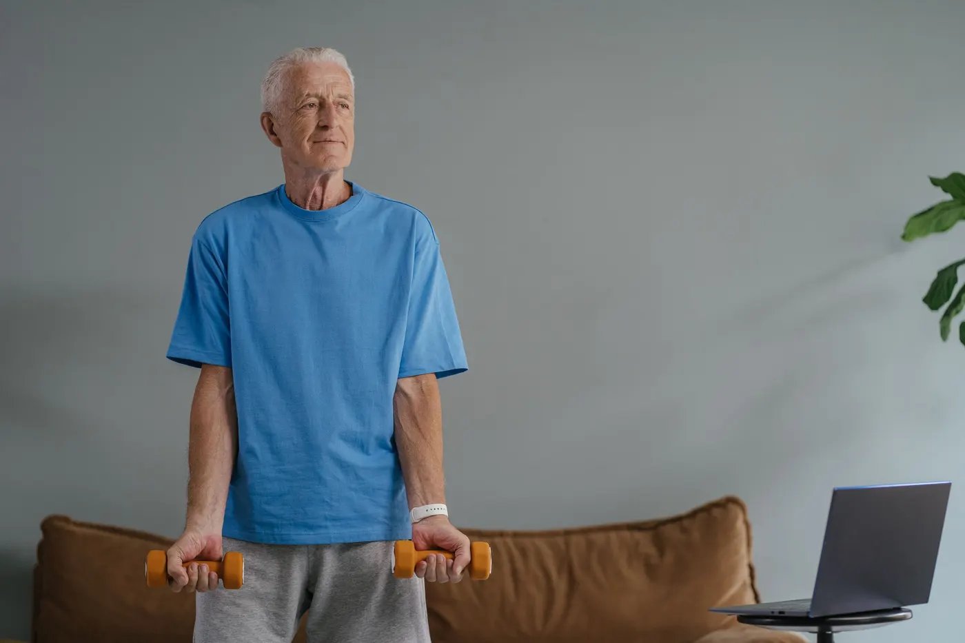 An older man doing dumbbell curls at home with a laptop open beside him