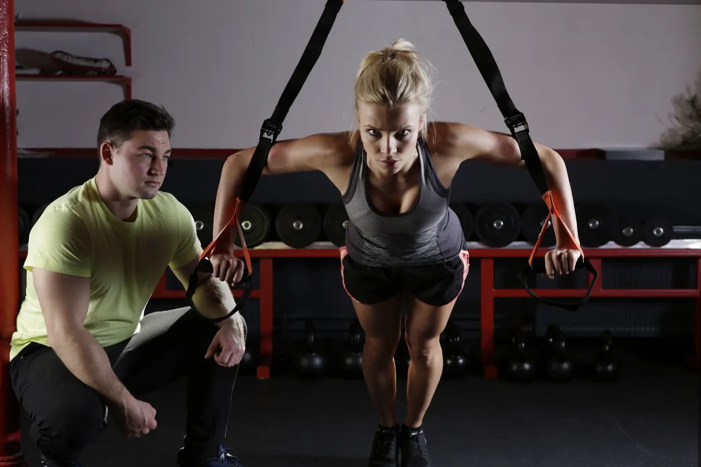 A trainer kneeling beside a woman and watching her form as she leans into suspension straps