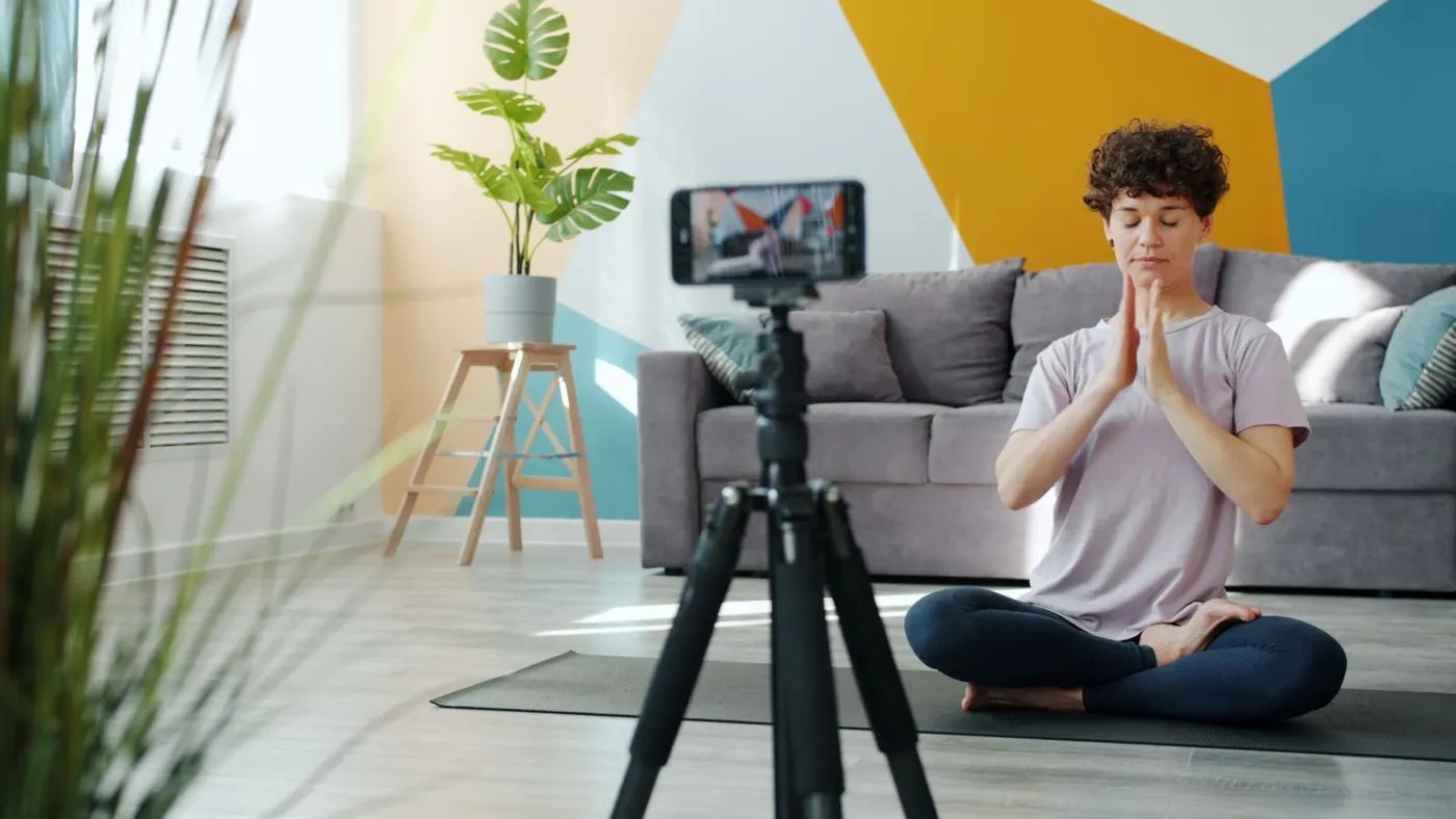 A woman on a mat in her living room facing a phone on a tripod that shows a live video
