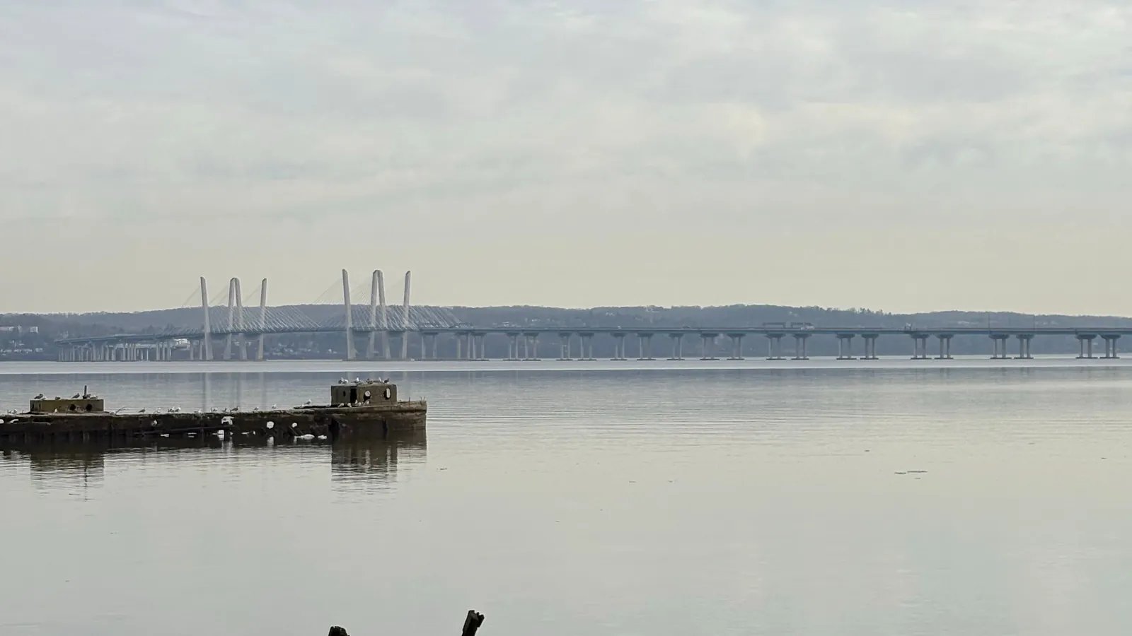 The Governor Mario M. Cuomo Bridge stretching across a calm Hudson River in daylight, with old pier pilings in the foreground